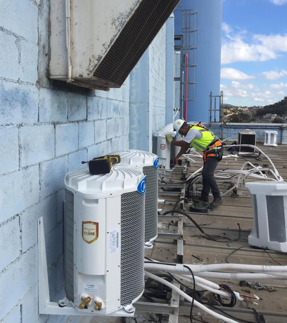 Técnico da Elcan Service, de capacete, colete e cinto de segurança, instalando condensadoras de ar condicionado na cobertura de um galpão